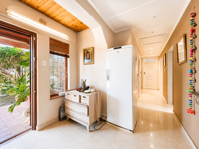 A bright hallway featuring tiled flooring, a timber-paneled ceiling, and recessed lighting, leading towards an interior doorway and an exterior patio entrance.