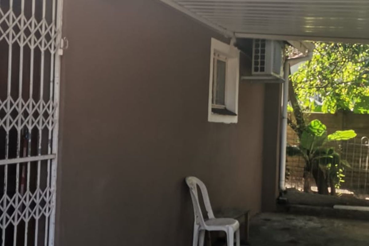 A covered outdoor patio area featuring a corrugated metal roof, security gate, and wall-mounted air conditioning unit, with concrete flooring.