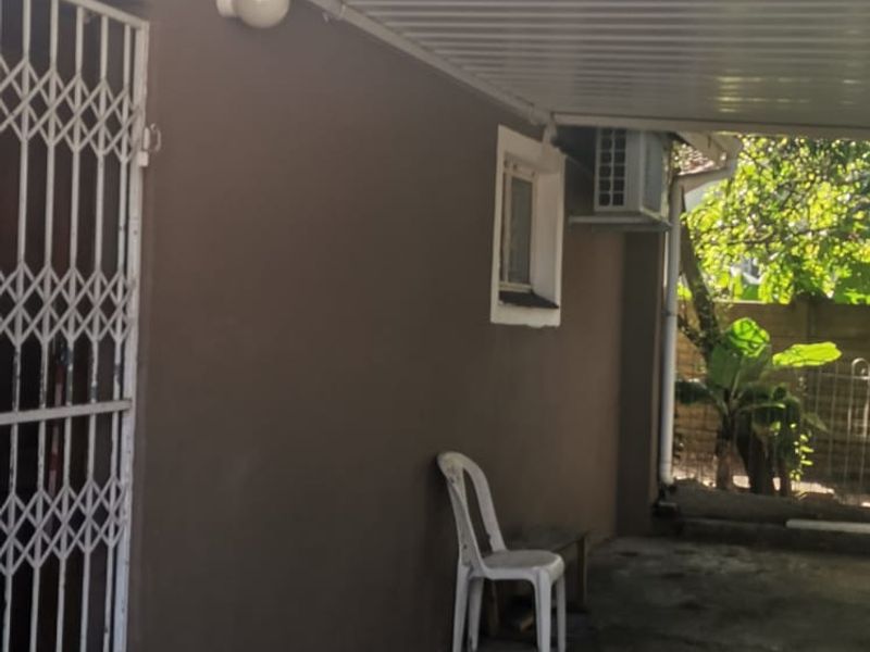 A covered outdoor patio area featuring a corrugated metal roof, security gate, and wall-mounted air conditioning unit, with concrete flooring.