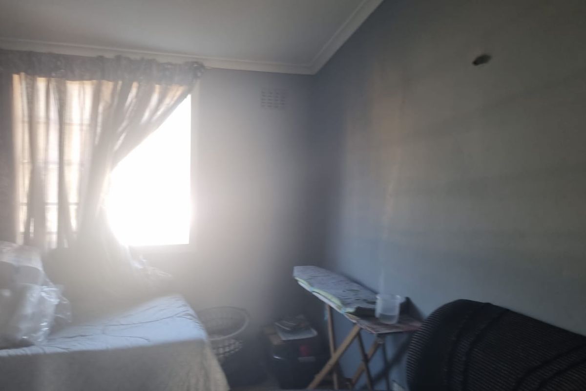 A bedroom featuring a window with curtains and wall-mounted ventilation, currently showing signs of wear and surface staining.