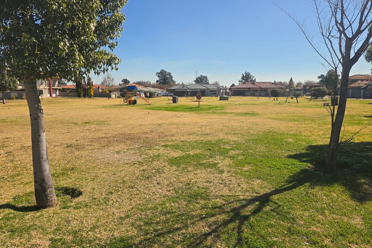 A spacious, open residential garden area featuring established trees, a grass lawn, and communal playground equipment in a suburban setting.