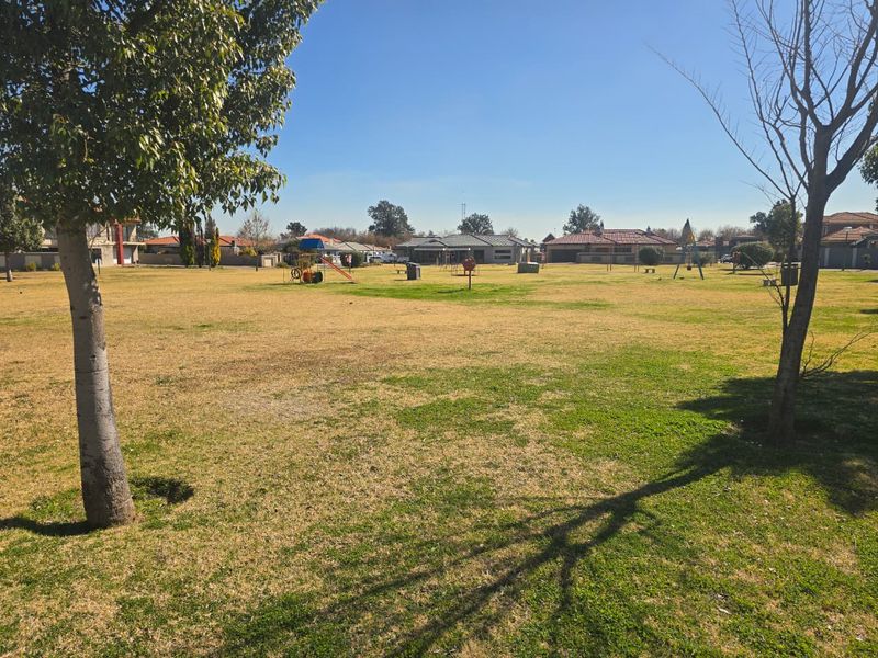 A spacious, open residential garden area featuring established trees, a grass lawn, and communal playground equipment in a suburban setting.
