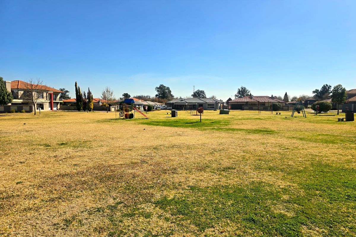 A large, open residential garden area featuring a grass lawn and communal playground equipment, surrounded by neighboring residential properties.