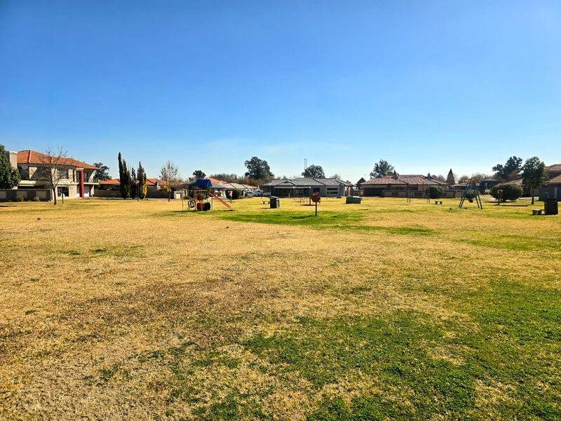 A large, open residential garden area featuring a grass lawn and communal playground equipment, surrounded by neighboring residential properties.