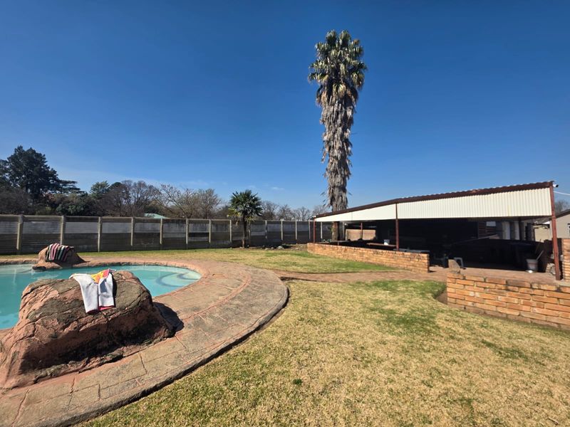Outdoor pool area featuring a paved surround, brick retaining walls, and a covered patio structure, set within a fenced garden.