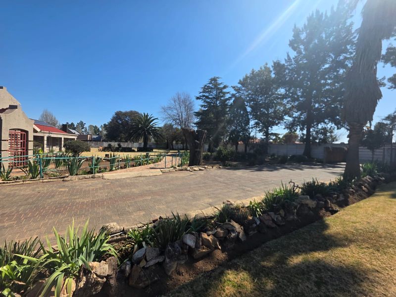A spacious front exterior view featuring a paved driveway, established garden with stone retaining walls, and mature trees under a clear blue sky.