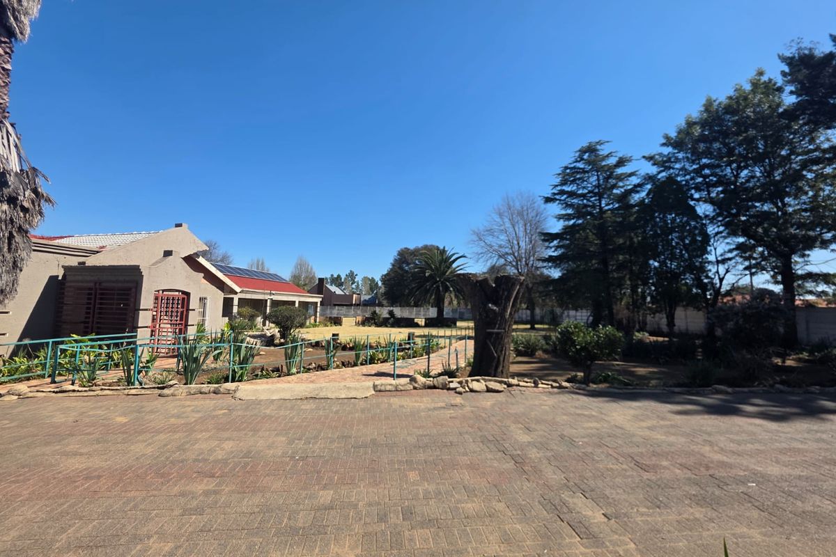 Spacious rear exterior view featuring a large paved driveway, established garden, and a residential building equipped with solar panels.