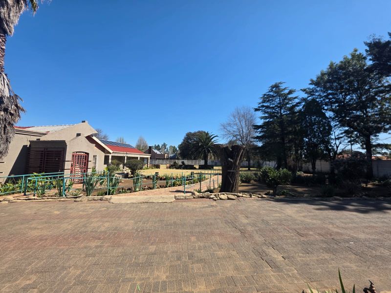 Spacious rear exterior view featuring a large paved driveway, established garden, and a residential building equipped with solar panels.