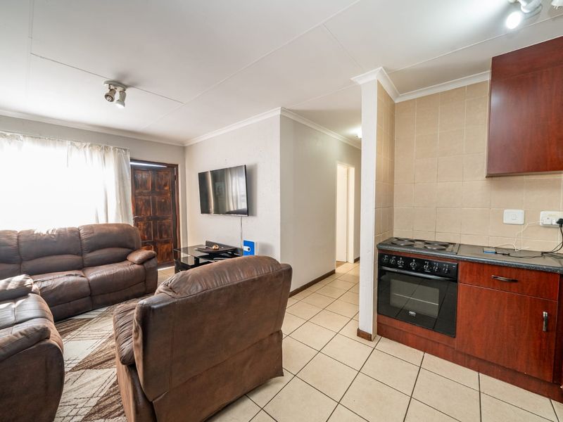 Open-plan living area featuring a compact kitchen with built-in cabinetry, electric stove, and oven, seamlessly flowing into the lounge space with tiled flooring.