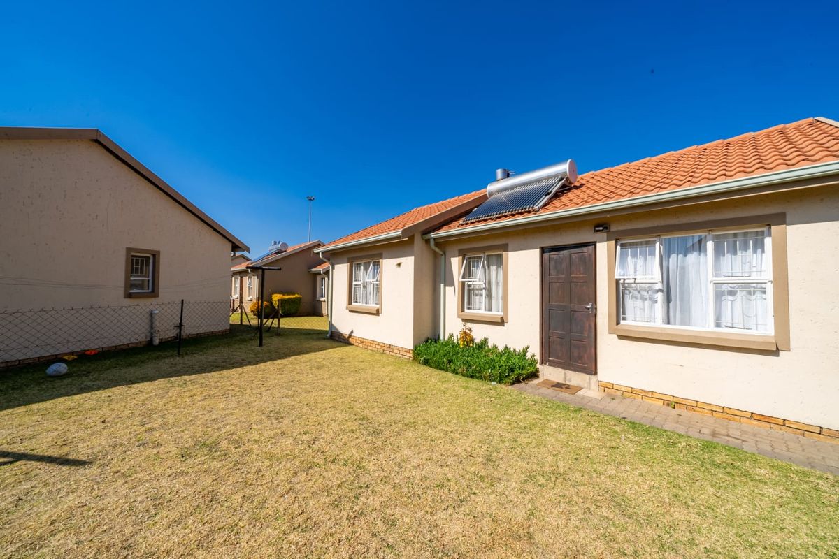 Rear view of the property featuring a lawn, paved walkway, and a solar water heating system installed on the tiled roof.