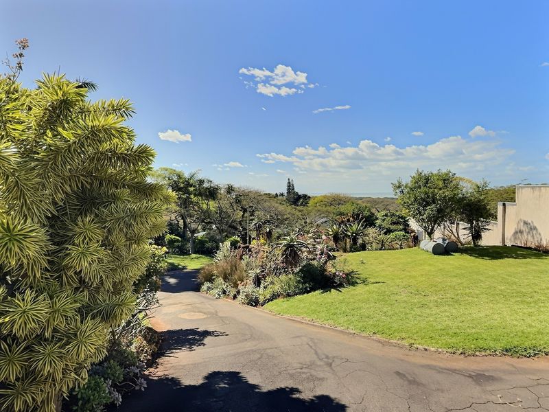 A well-maintained garden area featuring a paved driveway, lush lawn, and established landscaping with a view of the surrounding greenery.