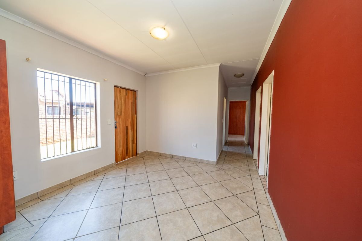 An empty living area featuring tiled flooring, a wooden door, and security bars on the window, leading into a hallway.