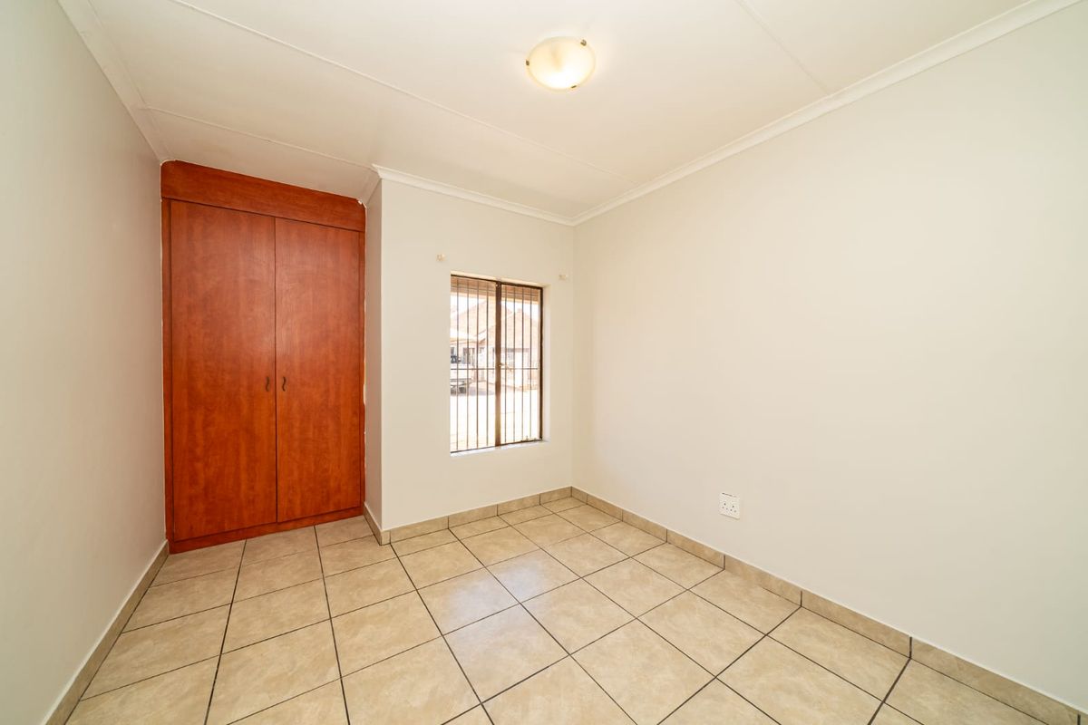 A clean, empty bedroom featuring neutral tiled flooring, a built-in wooden wardrobe, and a window fitted with security bars.