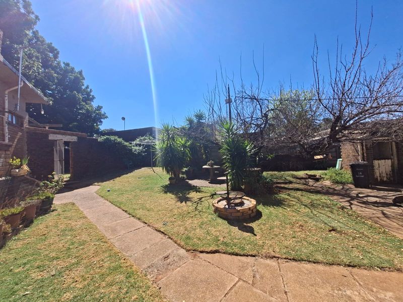 A spacious rear garden featuring a concrete pathway, established trees, and a built-in brick braai area, offering potential for outdoor landscaping.