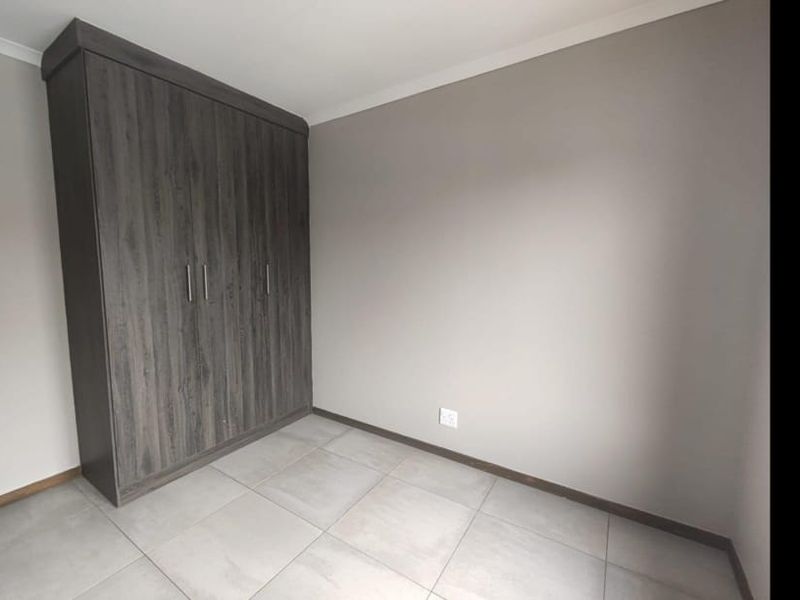 A clean, empty bedroom featuring neutral wall tones, large-format floor tiles, and a modern dark wood-grain built-in wardrobe.