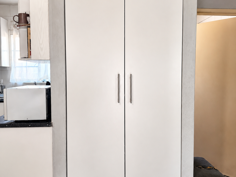 A clean kitchen area featuring a tall white built-in pantry cupboard, contrasting dark floor tiles, and functional cabinetry.
