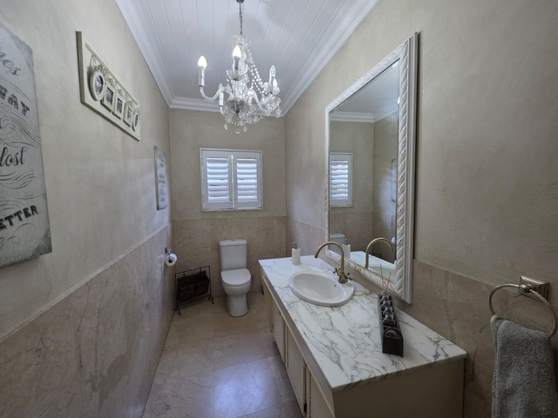 A well-appointed bathroom featuring a marble-topped vanity, brass fixtures, plantation shutters, and elegant chandelier lighting with classic tiled wainscoting.
