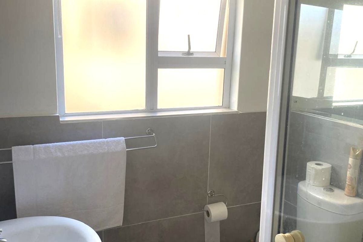 A functional bathroom featuring a pedestal basin, toilet, and a glass-enclosed shower with neutral wall tiling and a window for natural light.