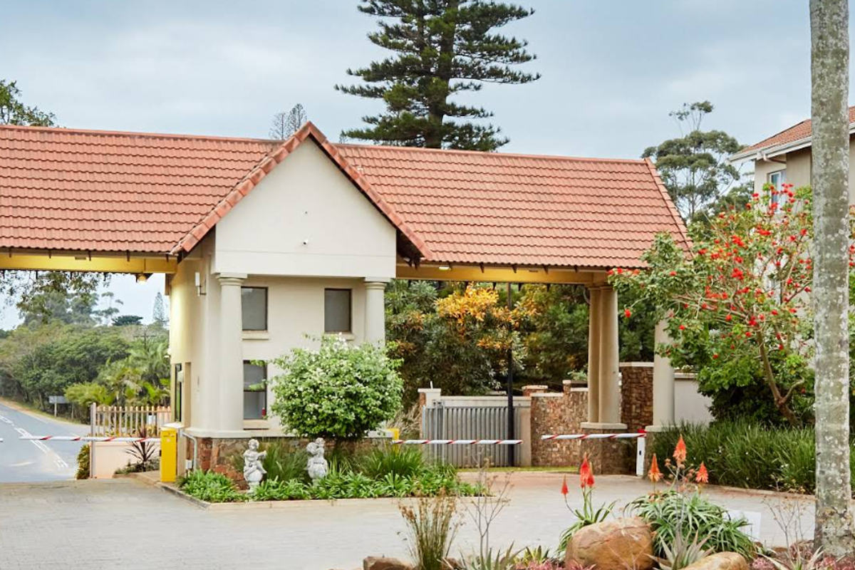 The entrance to the apartment complex features a secure gatehouse with a tiled roof, boom gate access, and paved driveway leading to the residential buildings.