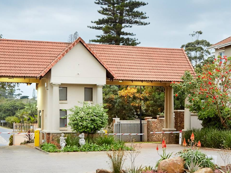 The entrance to the apartment complex features a secure gatehouse with a tiled roof, boom gate access, and paved driveway leading to the residential buildings.