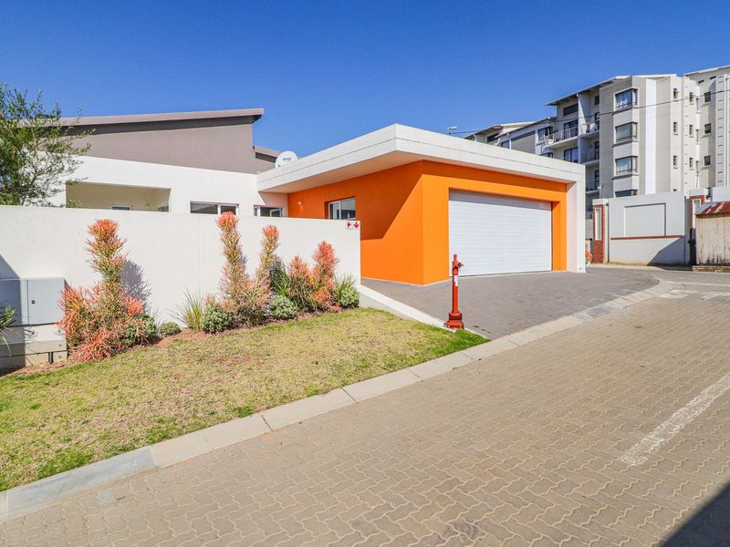 Front exterior view of a modern house featuring a bright orange garage facade, paved driveway, and a well-maintained boundary wall with landscaping.