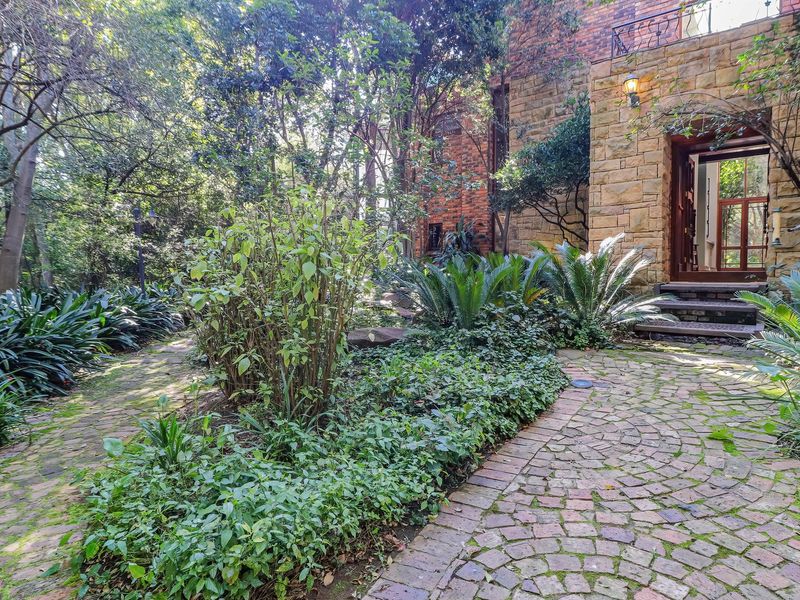 A charming entrance featuring a cobblestone pathway leading to a stone and brick facade, surrounded by lush, mature garden landscaping.