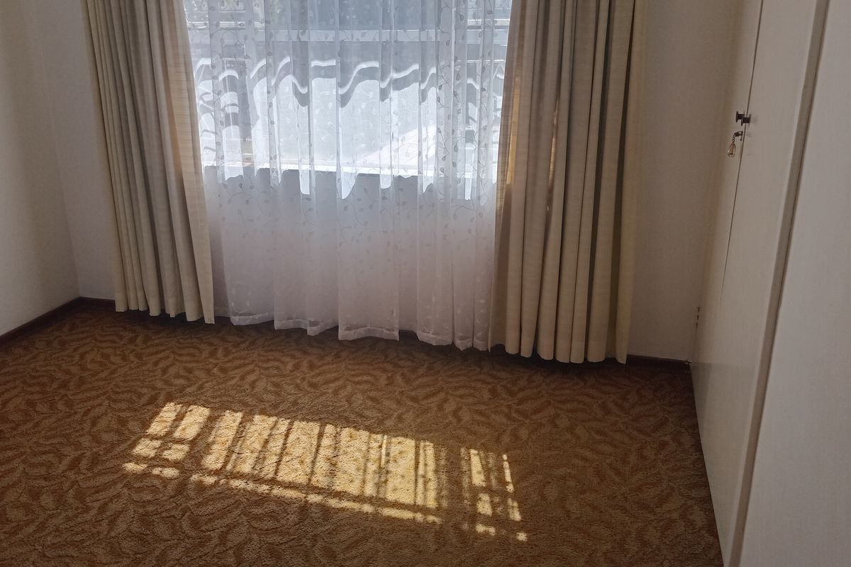 A vacant bedroom featuring patterned carpet flooring and a built-in wardrobe, illuminated by natural light through a window with curtains.