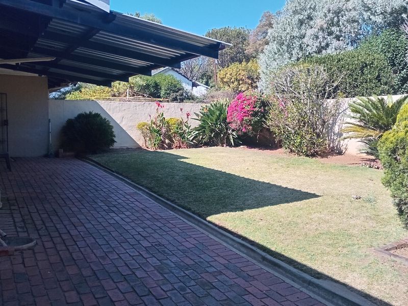 A rear garden area featuring a brick-paved patio covered by a corrugated metal roof, adjacent to a well-maintained lawn and boundary wall.
