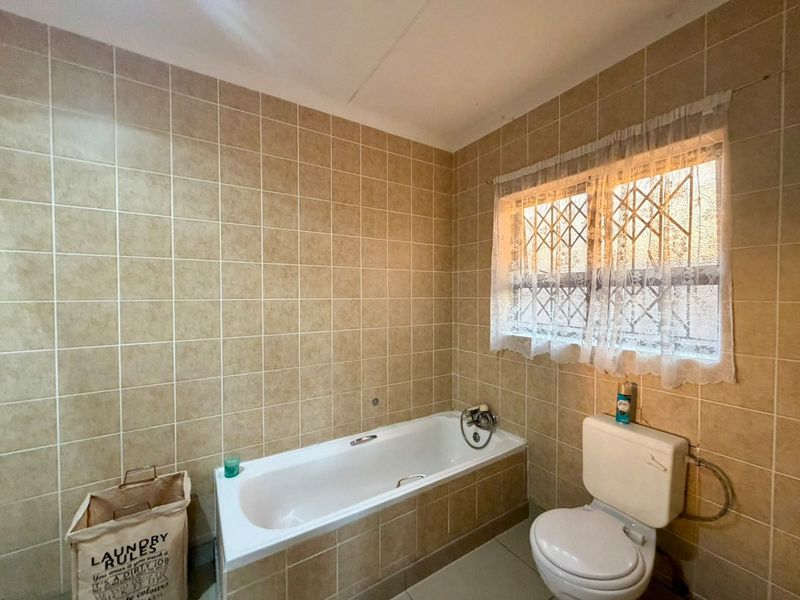 A dated bathroom featuring full-height ceramic wall tiling, a standard built-in bathtub, and a toilet, with security bars installed on the window.