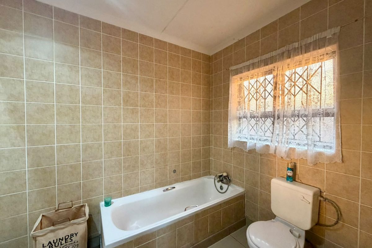 A dated bathroom featuring full-height ceramic wall tiling, a standard built-in bathtub, and a toilet, with security bars installed on the window.