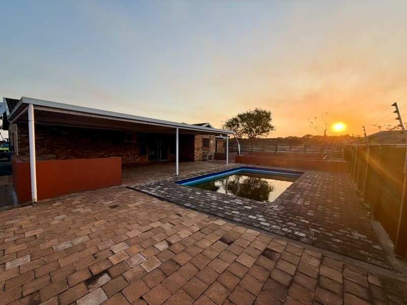 Outdoor entertainment area featuring a paved patio, covered veranda, and a swimming pool set against a sunset backdrop.