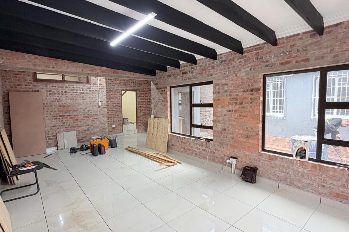 Spacious living area featuring exposed brick walls, dark timber ceiling beams, and large windows, currently undergoing renovation with tiled flooring throughout.