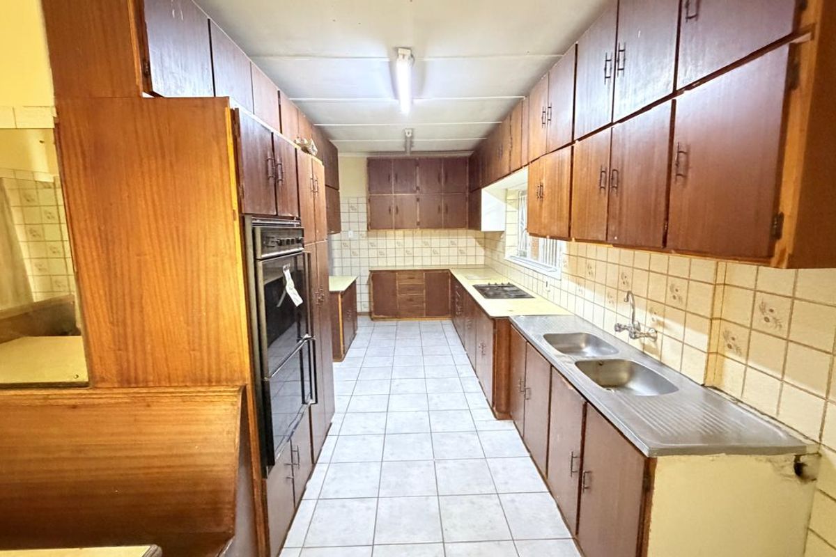 A spacious, dated kitchen featuring extensive wooden cabinetry, a built-in oven and hob, and a double stainless steel sink with tiled splashbacks.