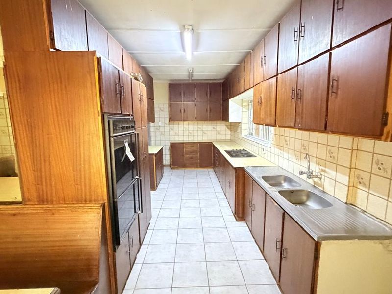 A spacious, dated kitchen featuring extensive wooden cabinetry, a built-in oven and hob, and a double stainless steel sink with tiled splashbacks.