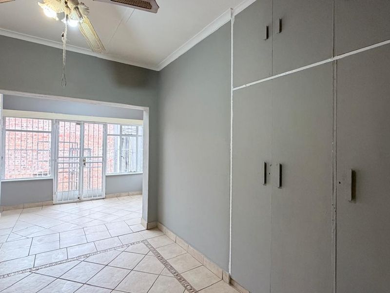 A spacious bedroom featuring neutral grey walls, built-in cupboards, tiled flooring, and a ceiling fan, with an adjoining sunroom area secured by metal gates.