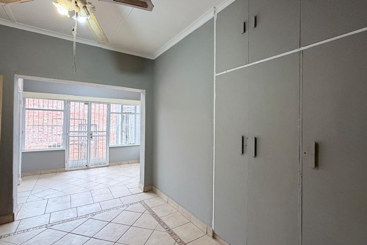 A spacious bedroom featuring neutral grey walls, built-in cupboards, tiled flooring, and a ceiling fan, with an adjoining sunroom area secured by metal gates.