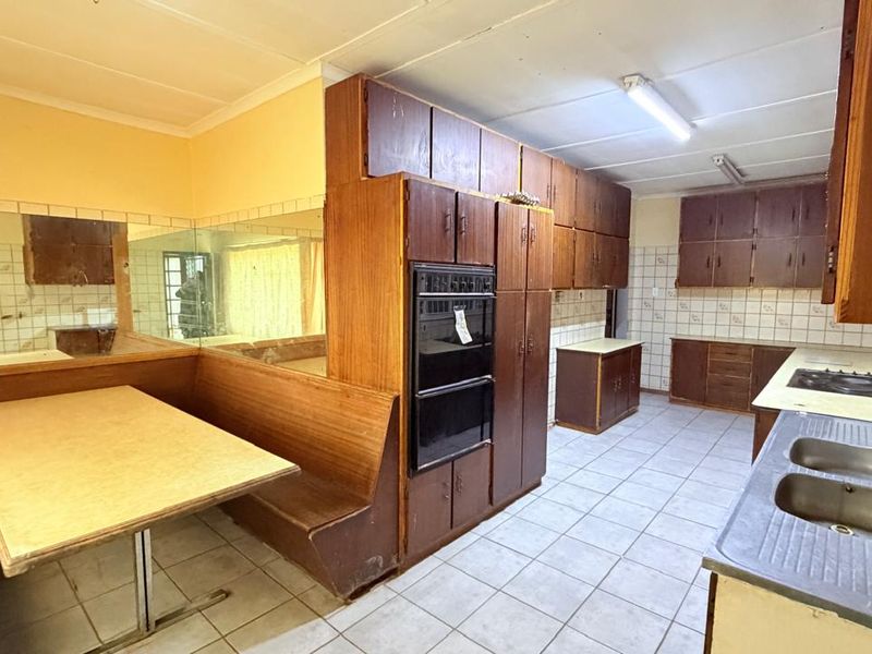 A dated kitchen featuring extensive wooden cabinetry, a built-in double oven, hob, double sink, and integrated bench seating with tiled finishes throughout.