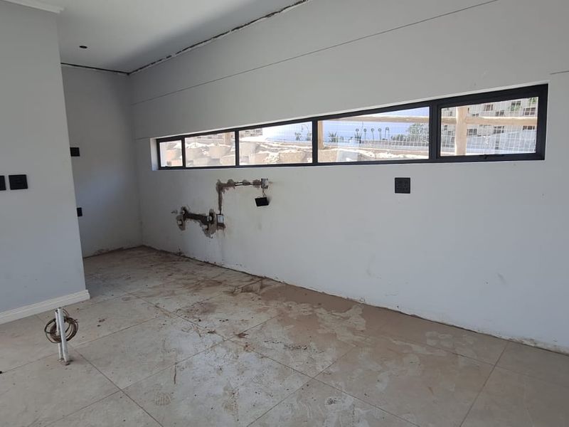 An unfinished kitchen space featuring tiled flooring, horizontal windows, and exposed plumbing rough-ins, ready for cabinetry and final finishes.