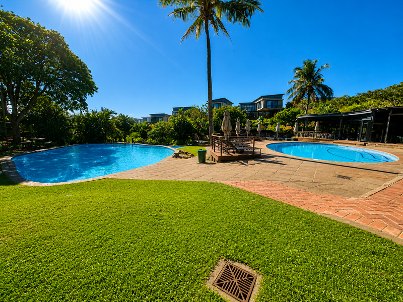 A well-maintained outdoor pool area featuring dual swimming pools, paved decking, brick pathways, and lush landscaped surroundings.