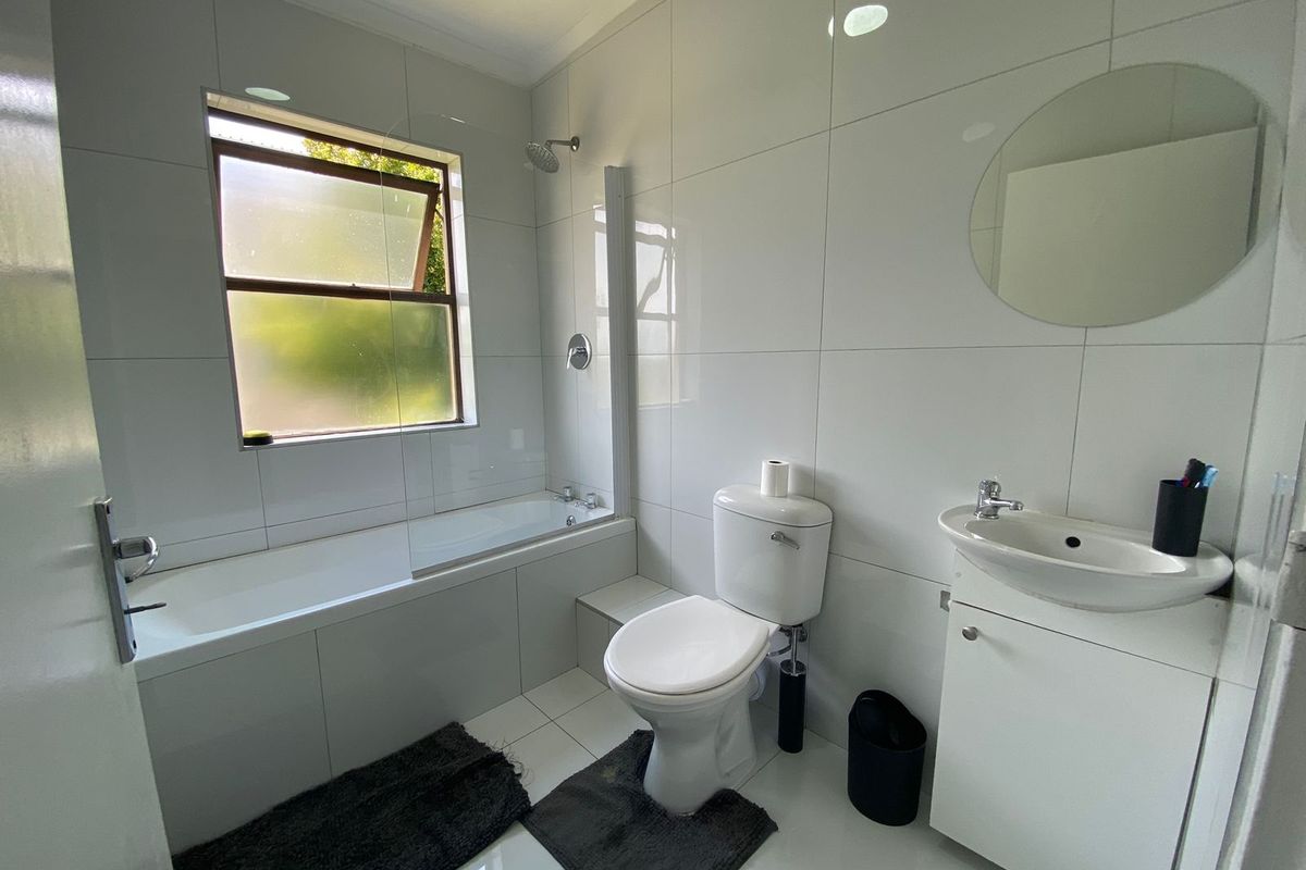 A clean, modern bathroom featuring white wall tiles, a built-in bathtub with a shower screen, a vanity basin, and a toilet.