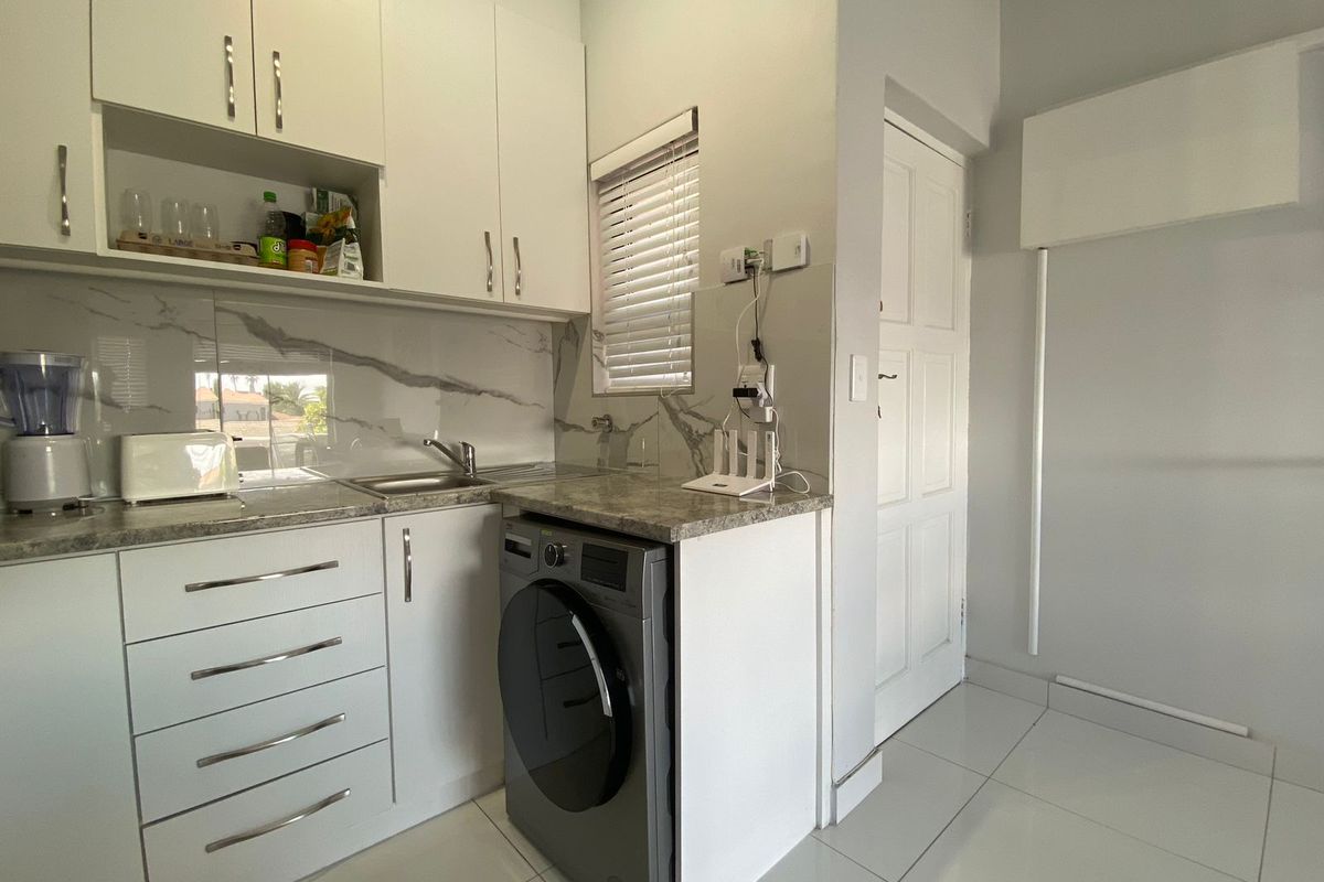 A compact, modern kitchen featuring white cabinetry, marble-effect backsplash, stone-look countertops, and integrated laundry appliance space with tiled flooring.