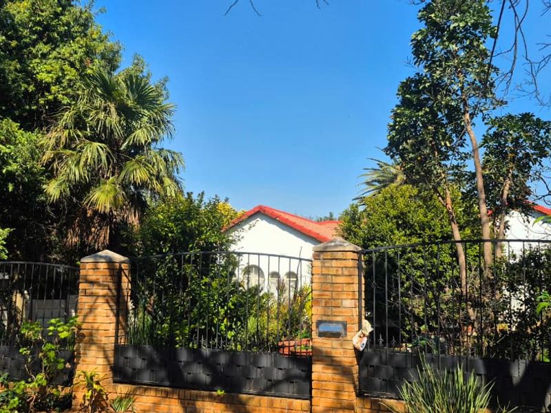 Front view of the property featuring a brick boundary wall with wrought iron fencing, mature garden landscaping, and a tiled roof structure.