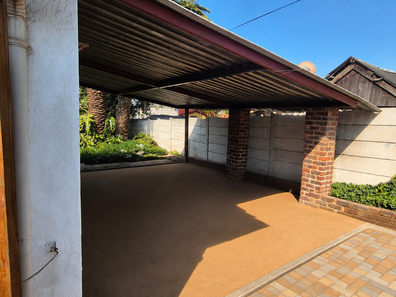 A covered carport area featuring brick support pillars, a corrugated metal roof, and a paved driveway, enclosed by a precast concrete wall.