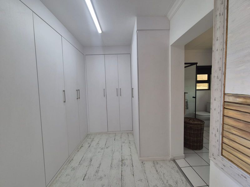 A clean, empty dressing area or bedroom featuring extensive white built-in cupboards and light-toned laminate flooring, with direct access to an adjoining bathroom.