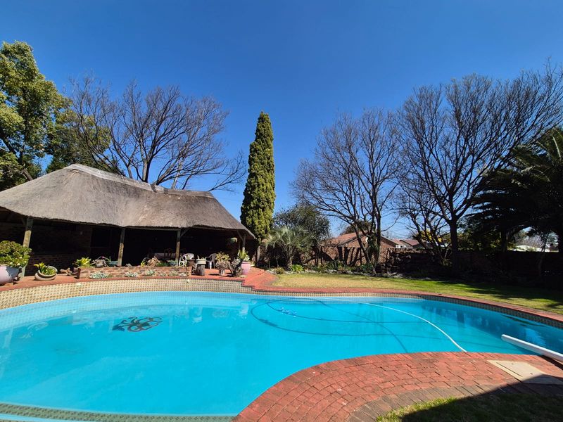 A backyard pool area featuring a swimming pool with brick coping and an adjacent thatched lapa structure set within a landscaped garden.