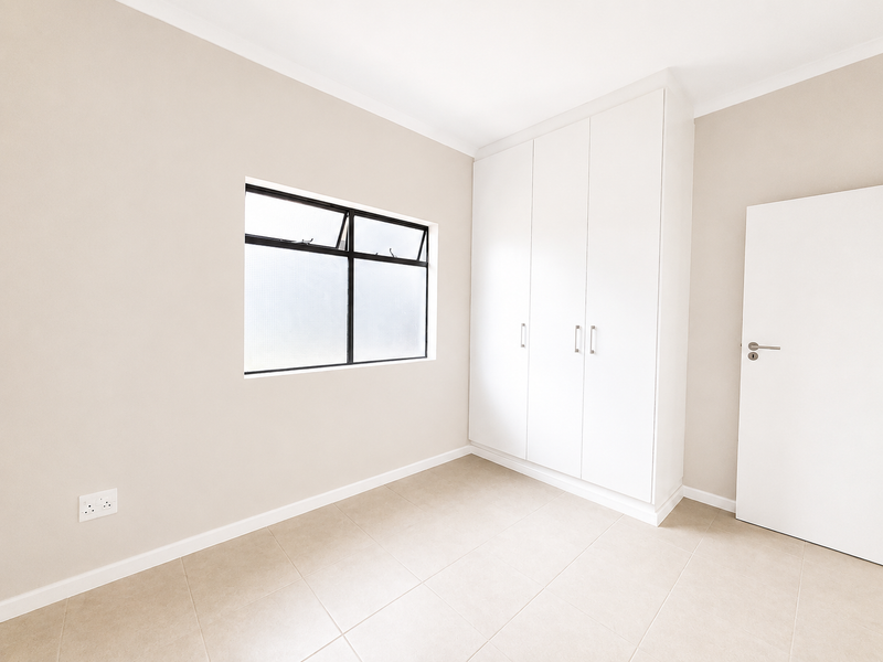 A clean, empty bedroom featuring neutral tiled flooring, built-in white cupboards, and a window providing natural light.
