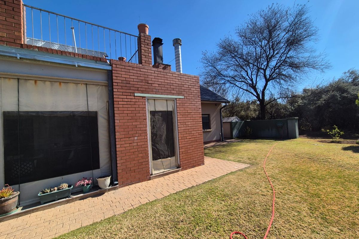 Rear exterior view of the house featuring a brick chimney, paved patio area, and a spacious lawn with metal balcony railing above.