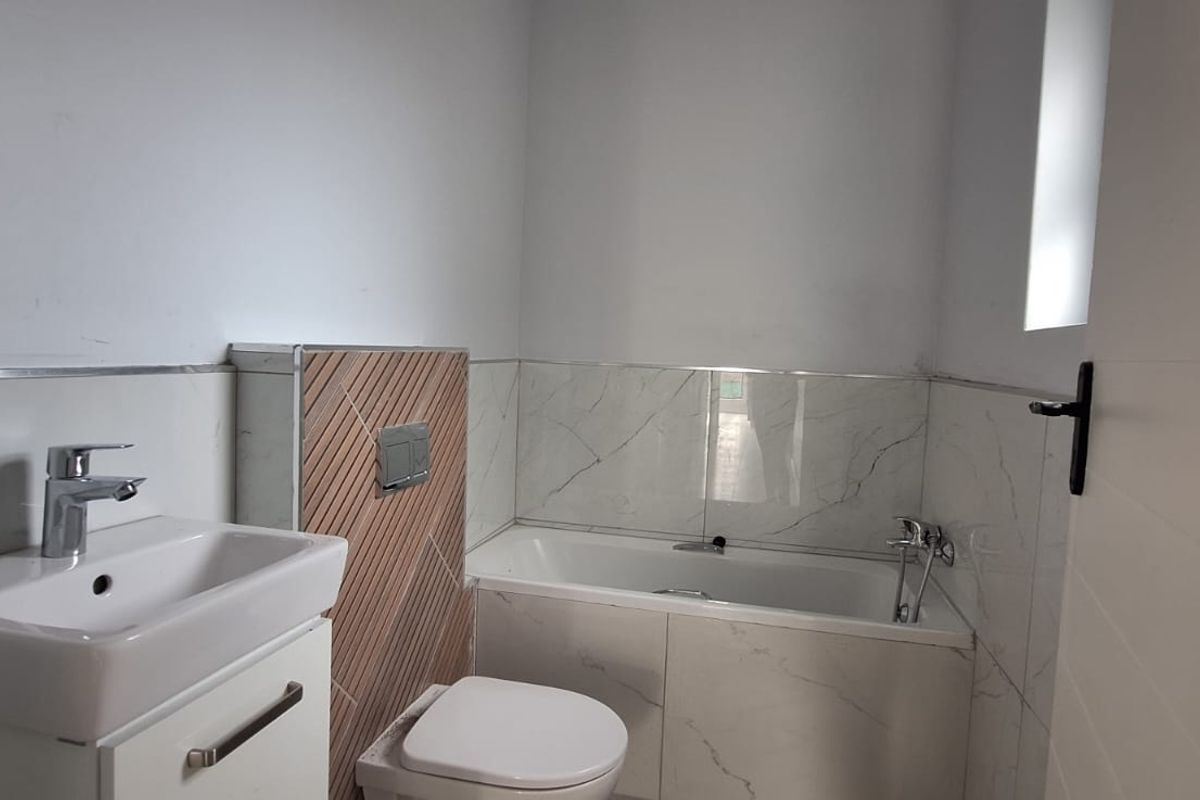 A modern, clean bathroom featuring a vanity basin, wall-hung toilet with concealed cistern, and a bathtub with marble-look wall tiling.