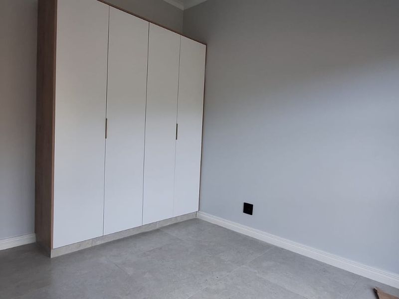 A clean, modern bedroom featuring neutral grey walls, large-format floor tiles, and a spacious built-in wardrobe with white cabinetry.
