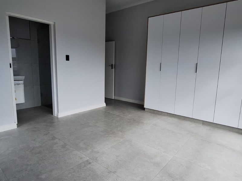 A bright, modern bedroom featuring large windows, neutral tiled flooring, and ample built-in cupboard space, with direct access to an en-suite bathroom.